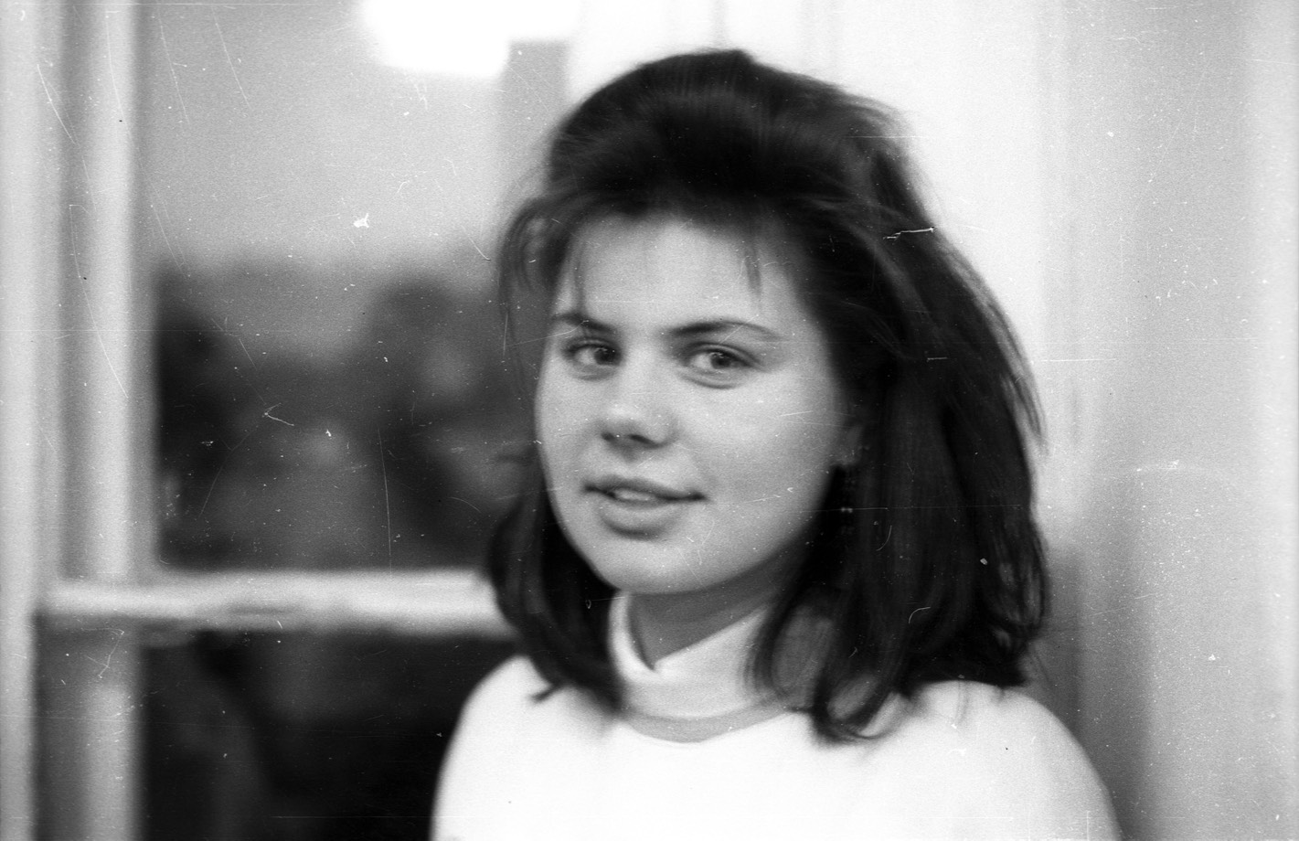 A scratched black-and-white portrait of a young girl in front of a window from the Fortepan collection