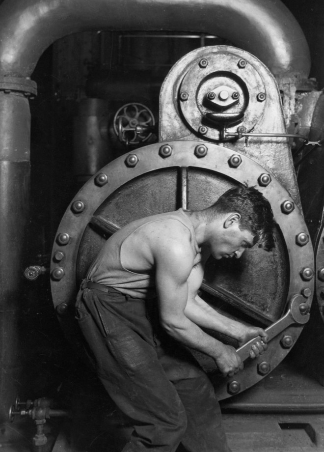 Black-and-white 1920 photograph of a mechanic bent over a steam pump with a wrench