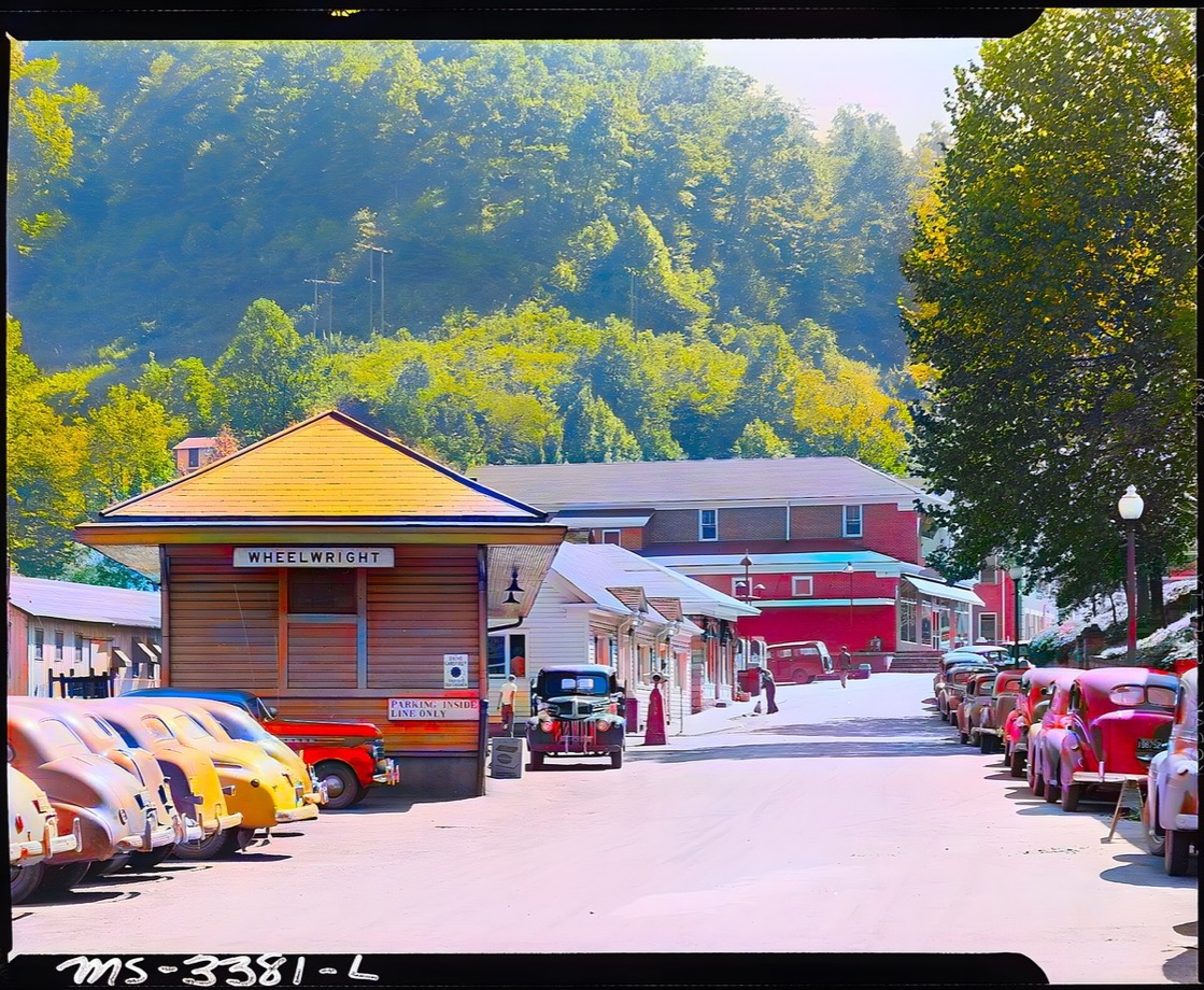 The same 1946 street colorized, with bright parked cars, green hillside trees and warm wooden buildings