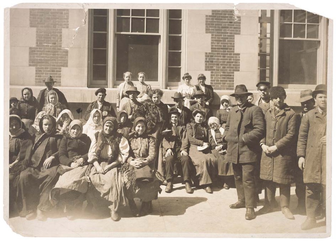 A black-and-white photograph of a group of immigrants outside a building on Ellis Island