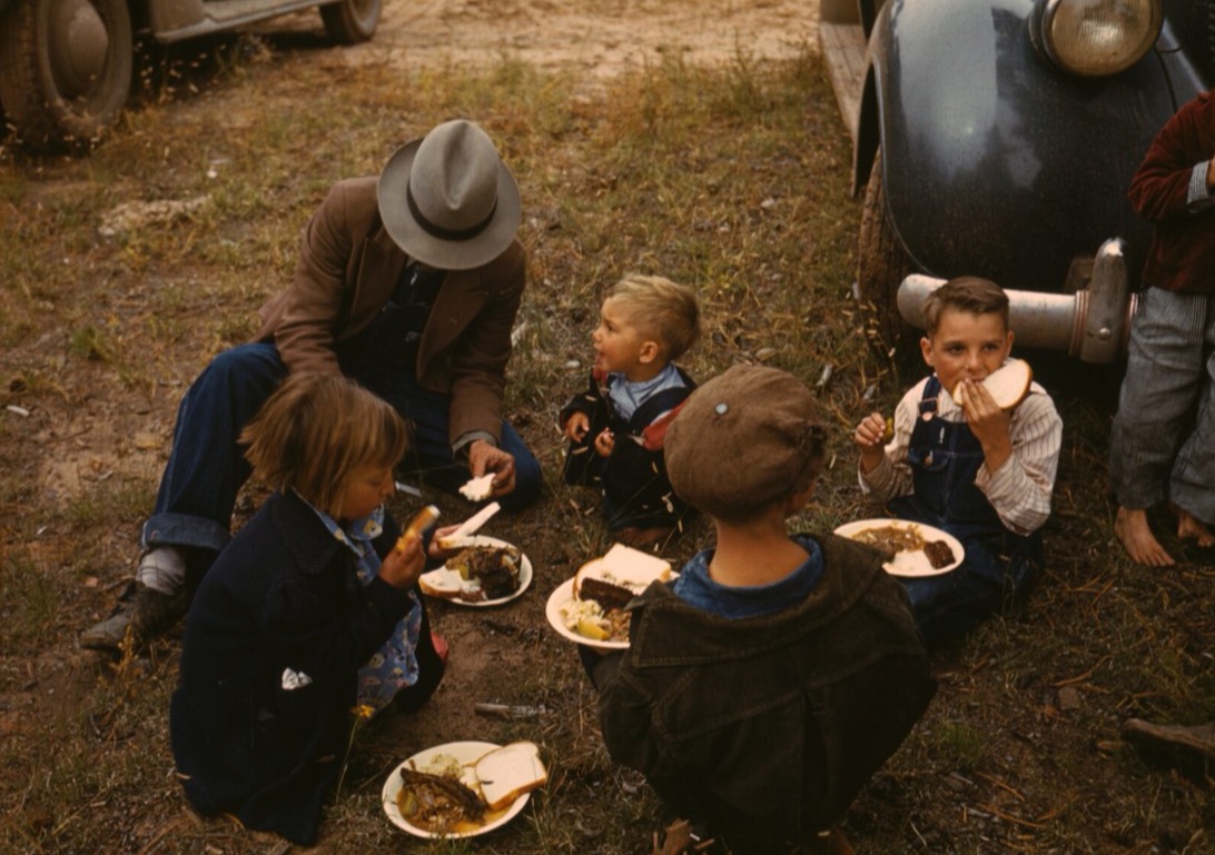 Black-and-white 1940 photograph of a father and five children eating barbecue on the grass beside parked cars