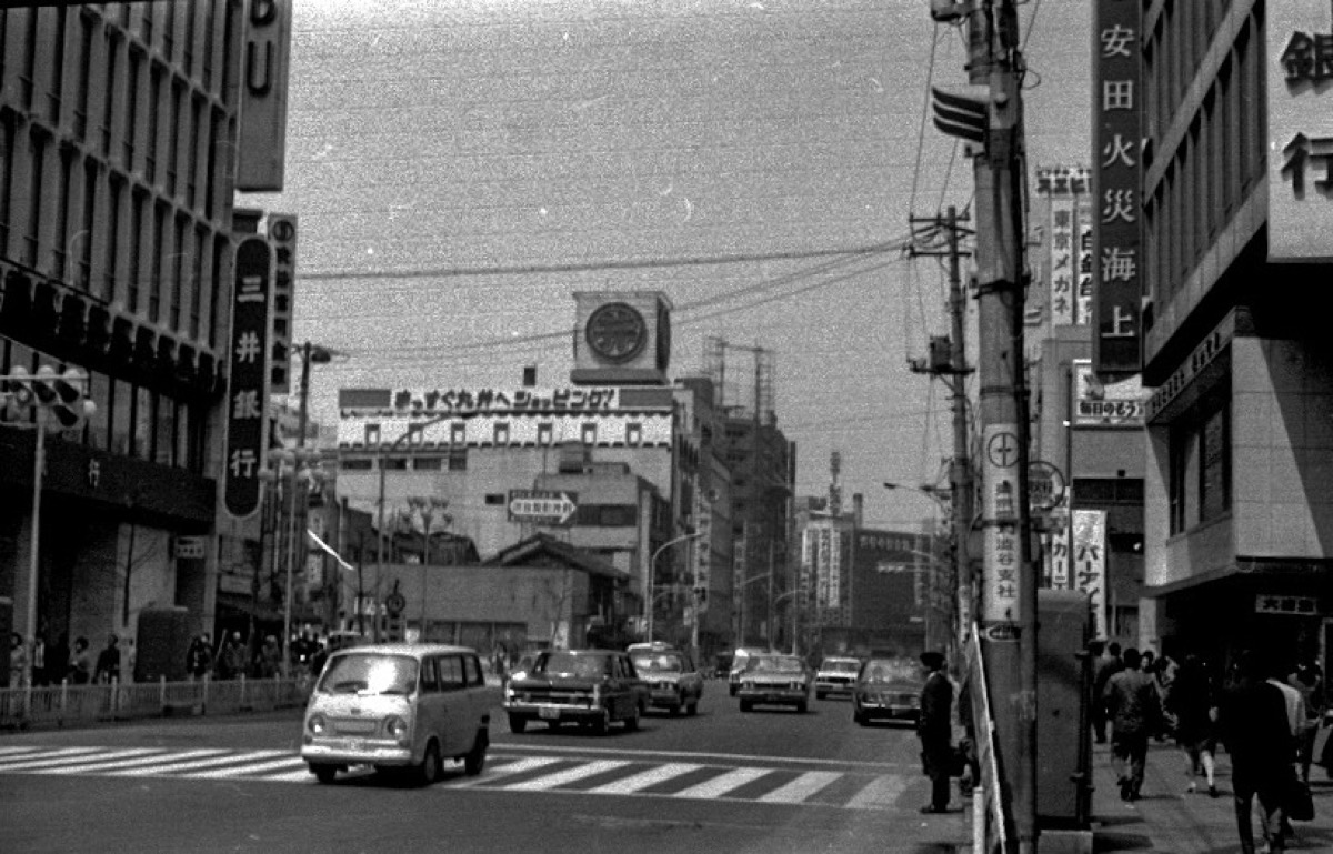 A black-and-white 1970 photograph of a Tokyo street with cars and shop signs